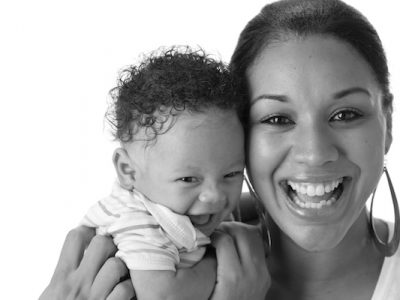 A head and shoulders image of a black mother with her one month old baby boy. Both are laughing.